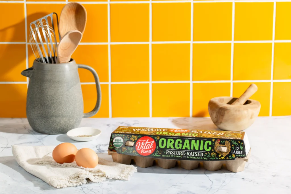 A carton of Vital Farms organic pasture-raised eggs on a counter with various kitchen tools against a vibrant yellow tiled backsplash.