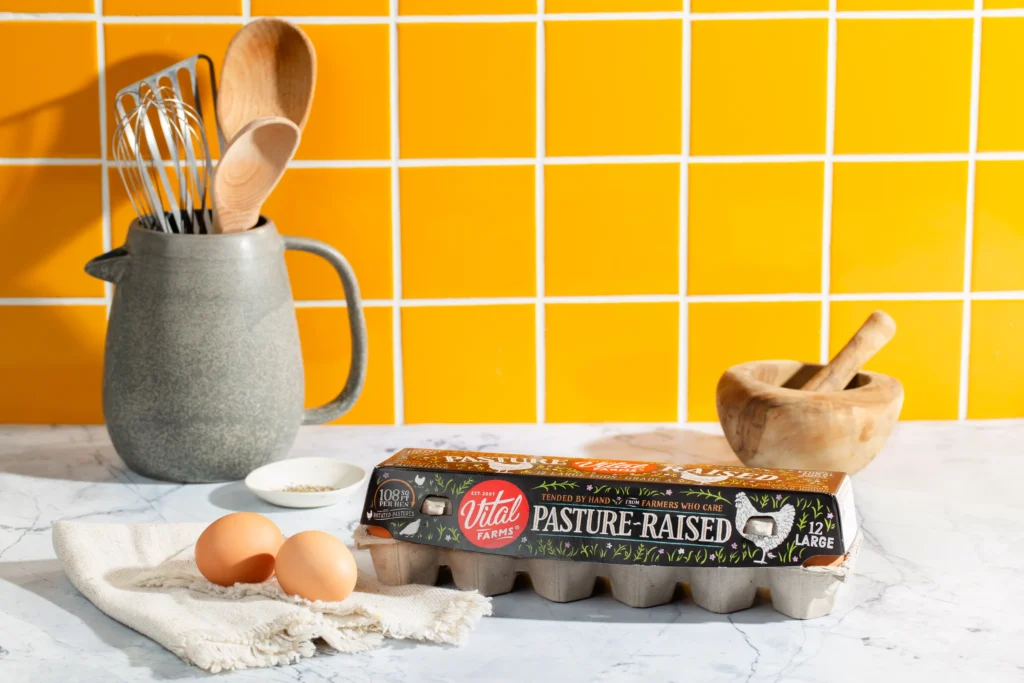 A carton of Vital Farms pasture-raised eggs on a counter with various kitchen tools against a vibrant yellow tiled backsplash.