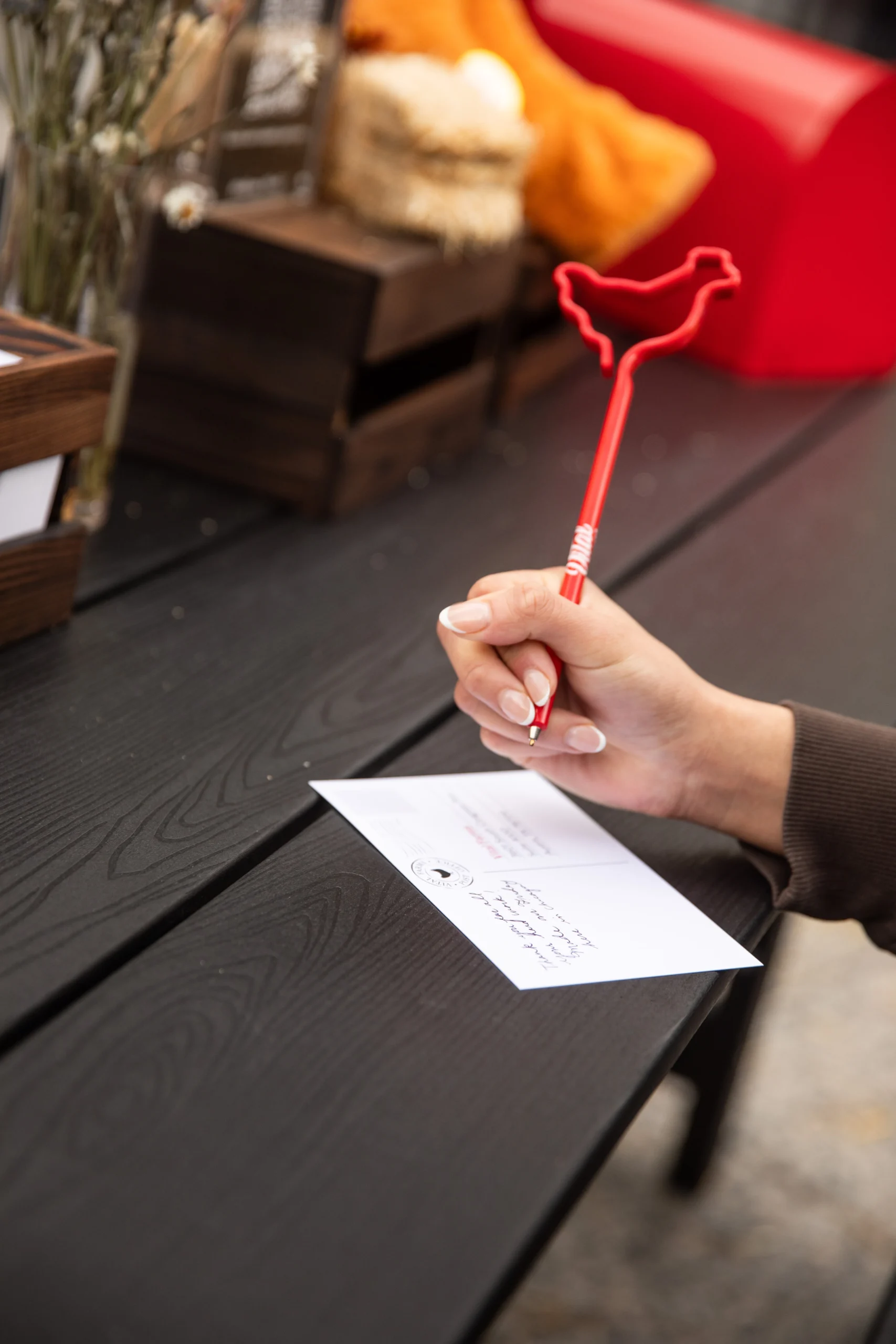 Person writes a note on a postcard on a dark brown surface, holding a red pen.