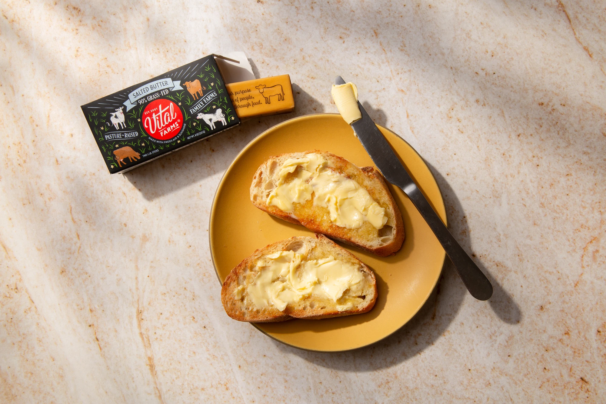 Buttered toast on a yellow plate with a knife and a carton of packaged butter.
