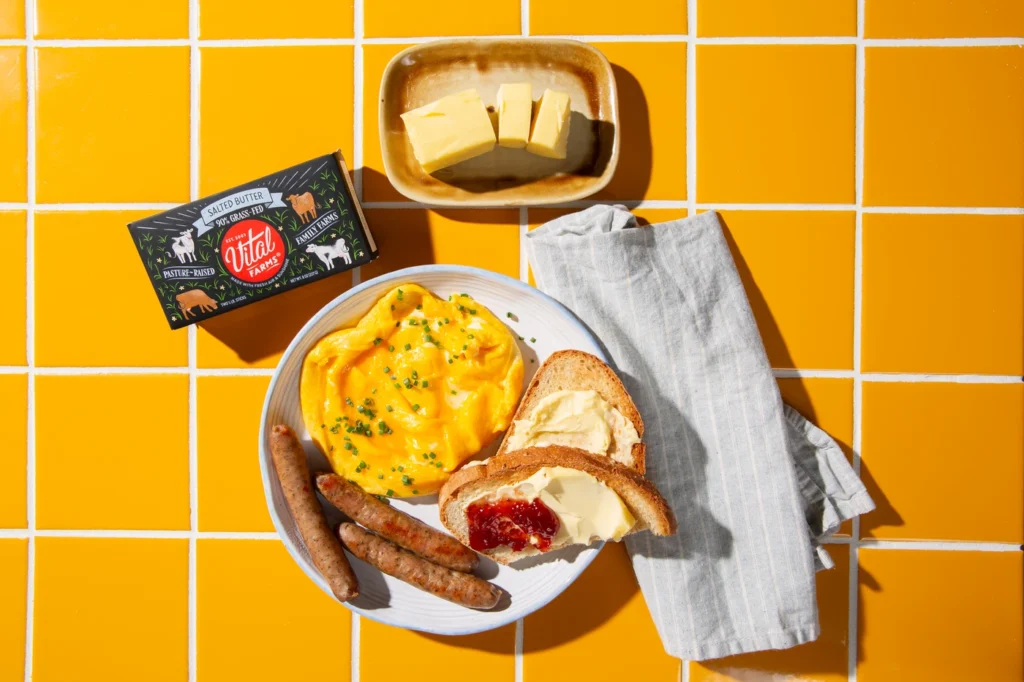 Plated breakfast of scrambled eggs, buttered toast with jam, and sausages on a yellow tiled countertop alongside a carton and tray of butter.